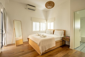 A bedroom with a wooden bed, white bedding, a bedside table, a mirror, and a window with blinds.