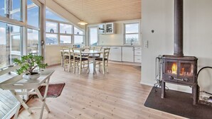 Amplia sala de estar con piso de madera, ventanas grandes, una mesa de comedor con sillas, una cocina con gabinetes blancos y un estufa de leña.