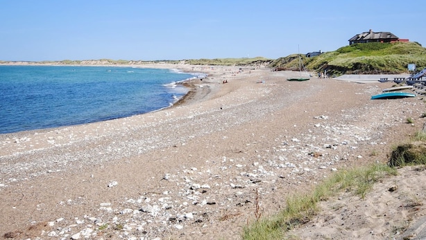 A pebble beach with a house on a hill, boats, and a clear sky.