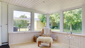 Ett rum med trägolv, en fåtölj med en fäll över ryggstöd och sits, en ljusstake och stora fönster med utsikt över grönområden utanför.