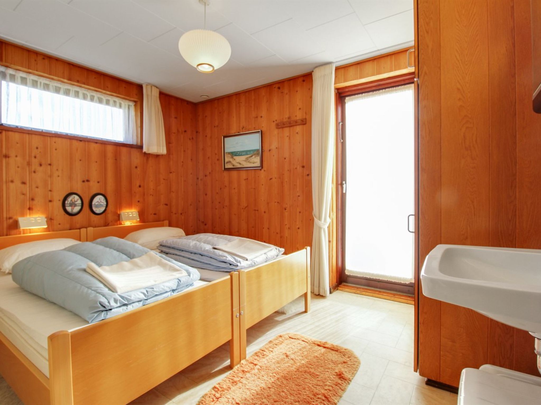 Una habitación con paneles de madera, dos camas, un pequeño cuadro y un baño con lavabo.
