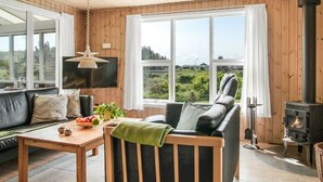 Un salon chaleureux avec un canapé, une table garnie de fruits et une plante, une cheminée et de grandes fenêtres donnant sur un paysage verdoyant.