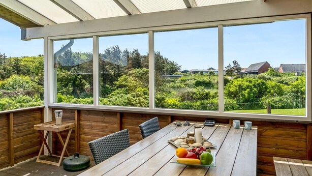 Une terrasse en bois avec une table dressée pour les repas, de grandes fenêtres offrant une vue sur un jardin luxuriant et des maisons, et une structure de plafond en bois.