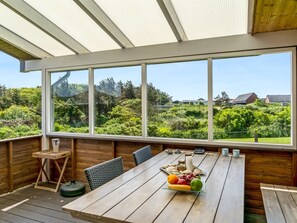 Eine Holzterrasse mit einem für das Essen gedecktem Tisch, große Fenster mit Blick auf einen üppigen Garten und Häuser, sowie ein hölzernes Deckenbauwerk.