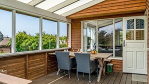 Une terrasse couverte avec une plateforme en bois, une table et des chaises, et une porte vitrée donnant sur un jardin.