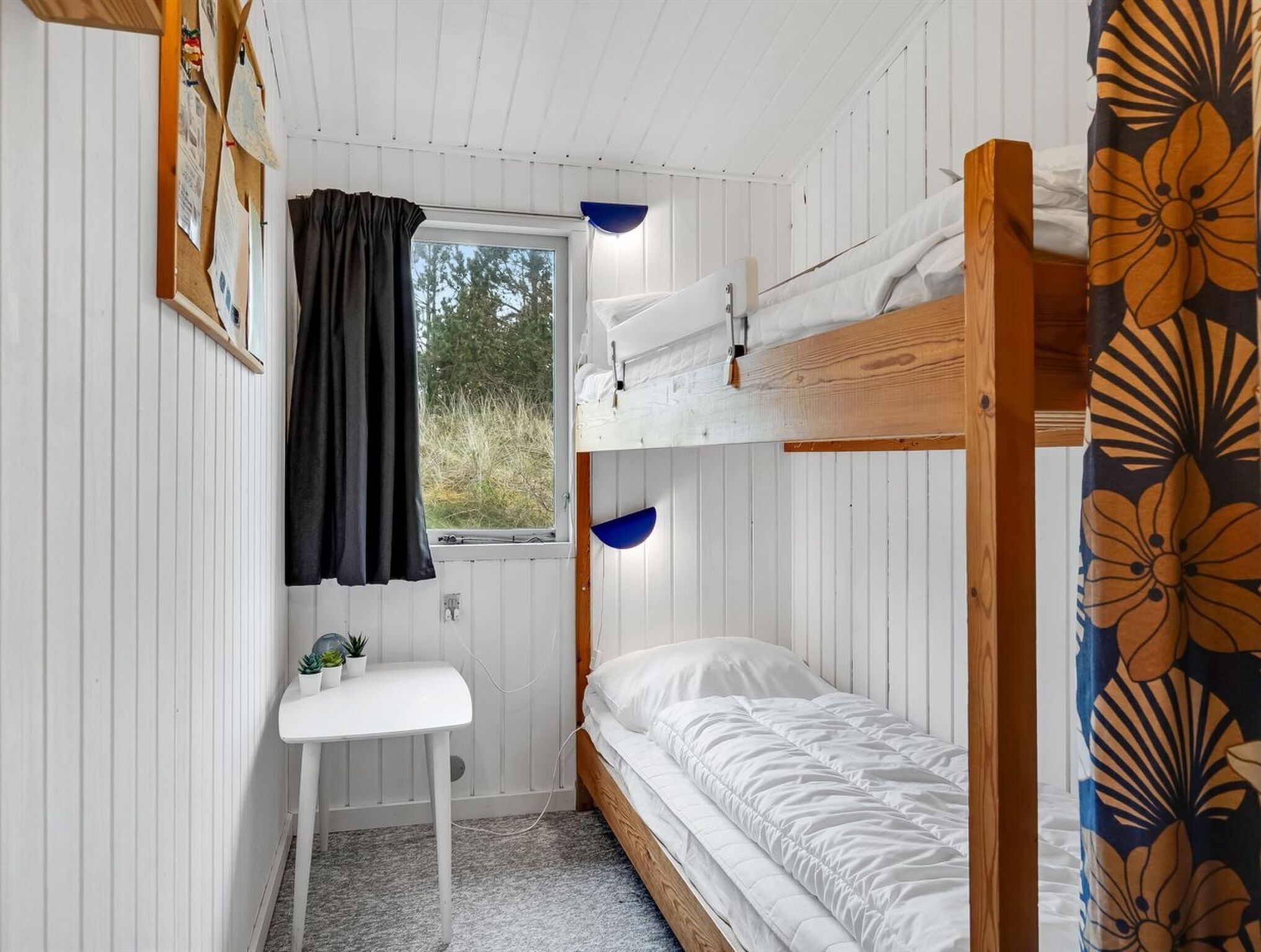 Een kleine kamer met een stapelbed, een bureau met een stoel, een raam met gordijnen en uitzicht op de omgeving.