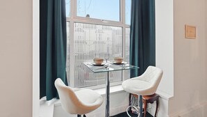 Una habitación de esquina con ventana, dos sillas blancas, una mesita de cristal y un radiador.