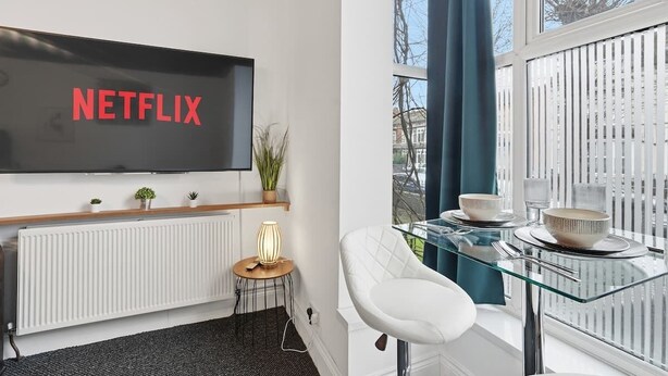 Una sala de estar moderna con un televisor de pantalla plana grande que muestra 'NETFLIX', una mesa de comedor de vidrio con sillas blancas, un radiador y una pequeña mesita de noche con una lámpara.