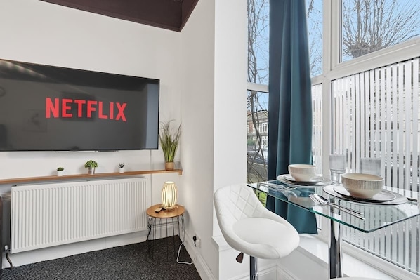 A modern living room with a large flat-screen TV displaying 'NETFLIX,' a glass dining table with white chairs, a radiator, and a small side table with a lamp.