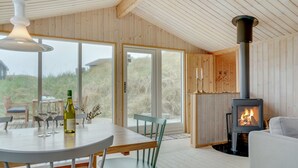 A cozy room with a wooden interior, a large window, a table with chairs, a sofa, and a wood-burning stove.