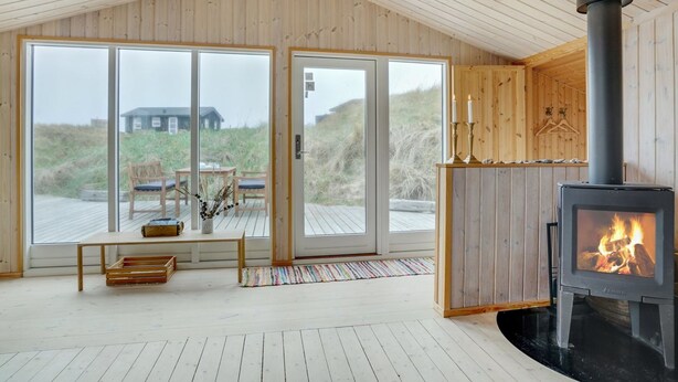 A cozy cabin interior with a large wooden ceiling, a fireplace, and a view of the beach through large glass doors.