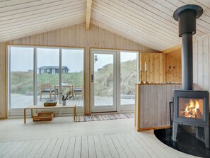 A cozy cabin interior with a large wooden ceiling, a fireplace, and a view of the beach through large glass doors.