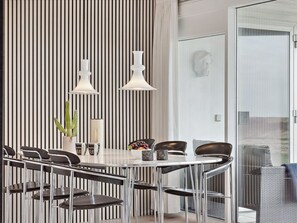 Un espace repas moderne avec une table blanche, des chaises noires, deux suspensions et un mur à rayures.