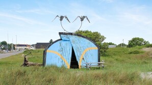 En rustik, vejrbidt blå bygning med en buet indgang og to metalkulpturer, der ligner insekter, øverst.