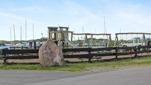 En legeplads med rutsjebane og klatrestrukturer, en stor klippe med udskæringer, samt en parkeringsplads med både og køretøjer.