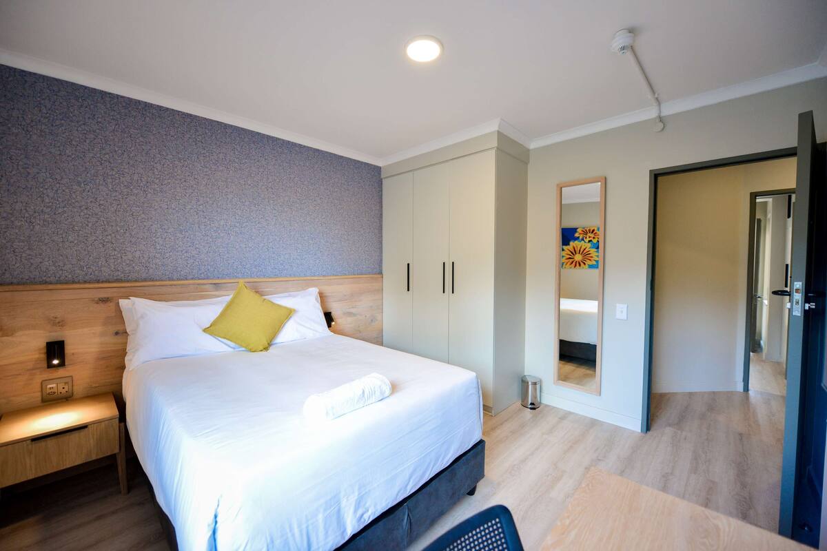 A modern hotel room with a large bed, a bedside table, a wardrobe, and a mirror.