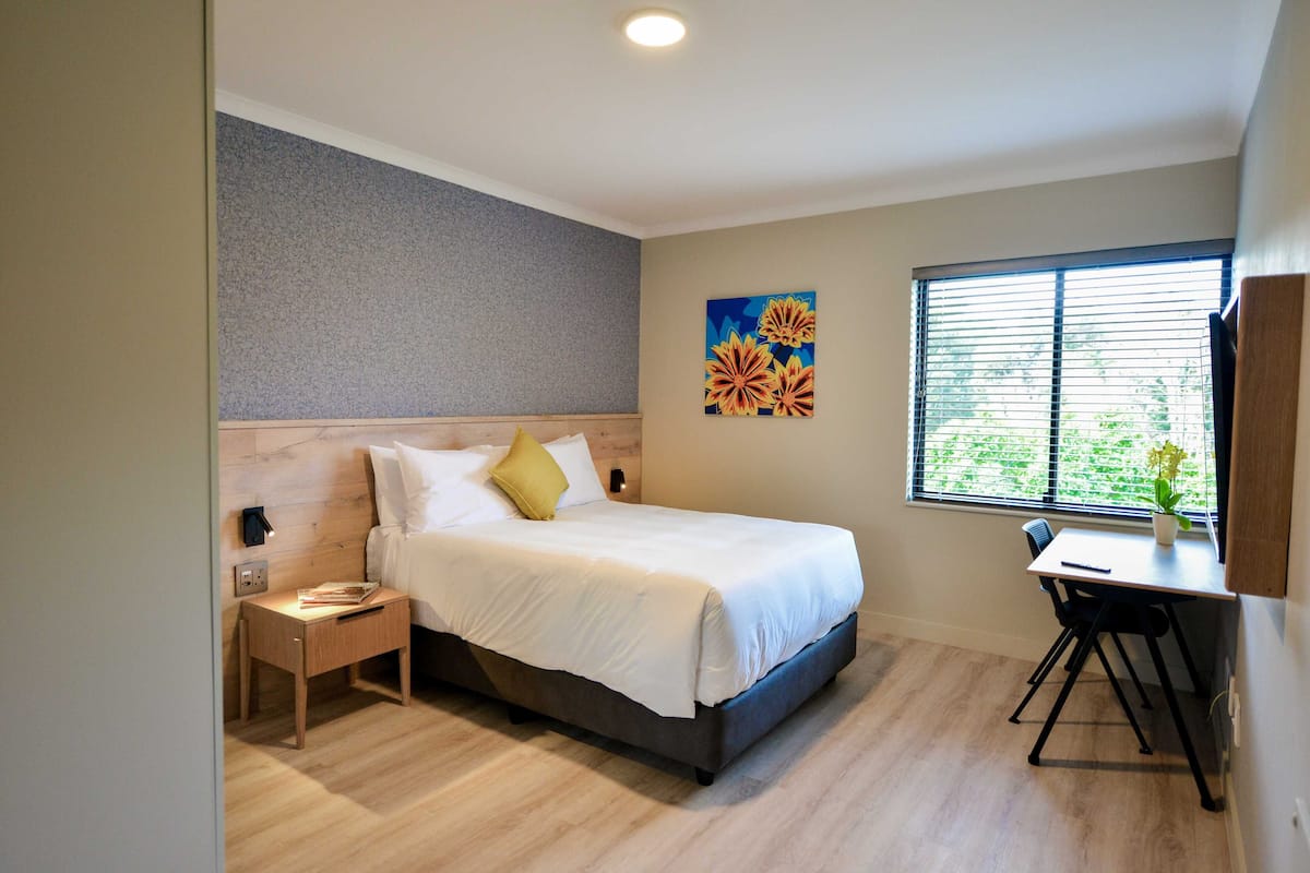 A modern hotel room with a bed, bedside table, desk, and window with blinds.