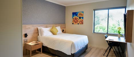 A modern hotel room with a bed, bedside table, desk, and window with blinds.