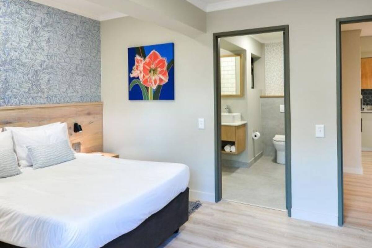 A bedroom with a bed, a nightstand, a painting, and a view into the bathroom and kitchen.