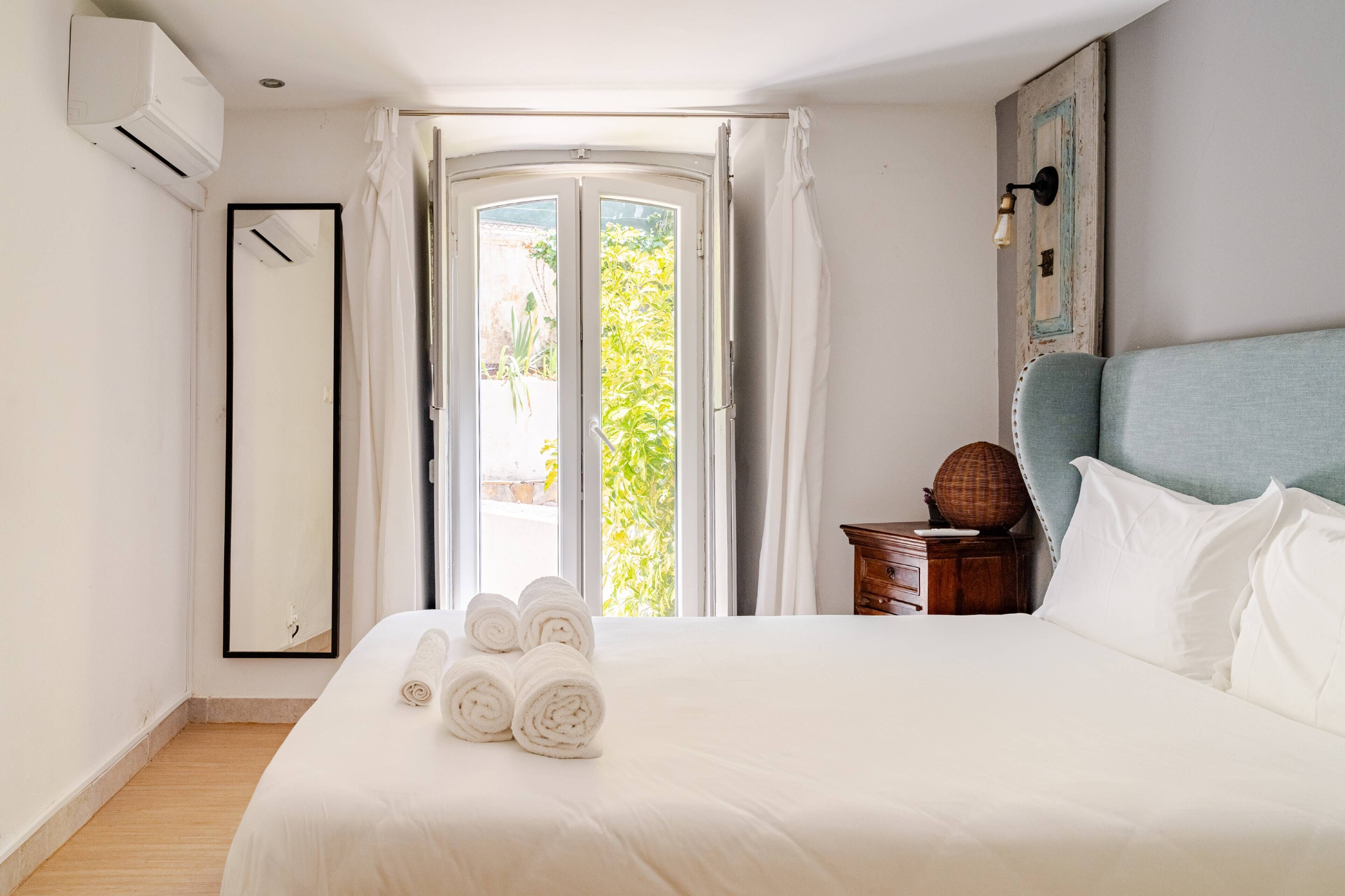 A hotel room with a bed, a nightstand, a mirror, and an air conditioning unit.