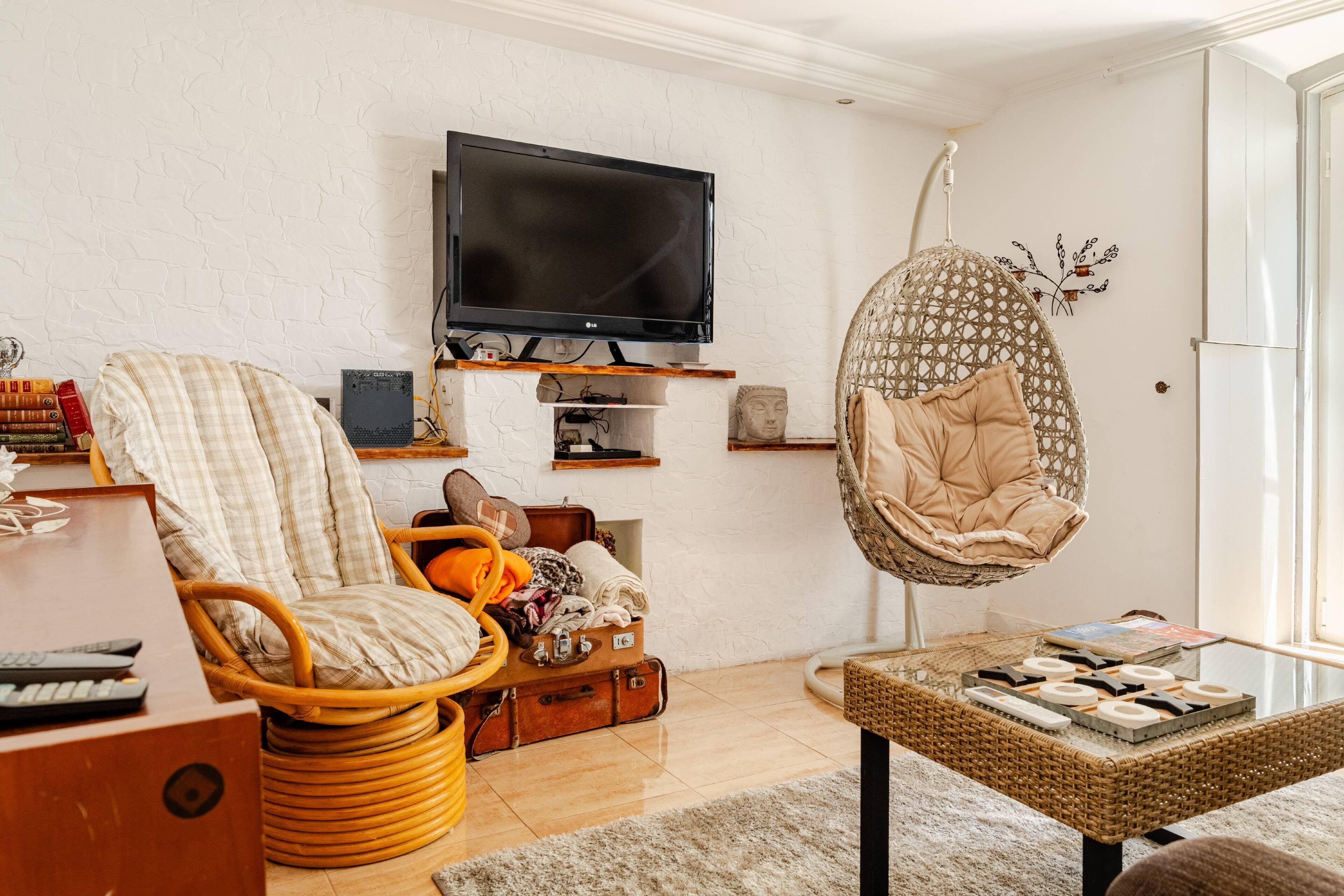 Una sala de estar con televisor, muebles de mimbre, una silla colgante y una estantería.