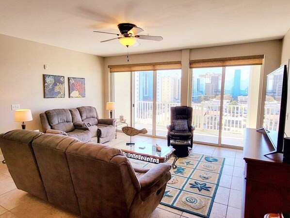 A living room with a sofa, armchair, and a coffee table. There is a ceiling fan, a TV, and a view of buildings through large windows.