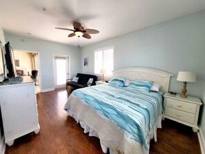 A bedroom with a large bed, a ceiling fan, a dresser, and a nightstand with a lamp.
