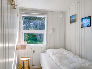 Ein kleines Zimmer mit einem Einzelbett, weißen Holzwänden, einem Fenster mit Blick auf Bäume und zwei gerahmten Bildern an der Wand.