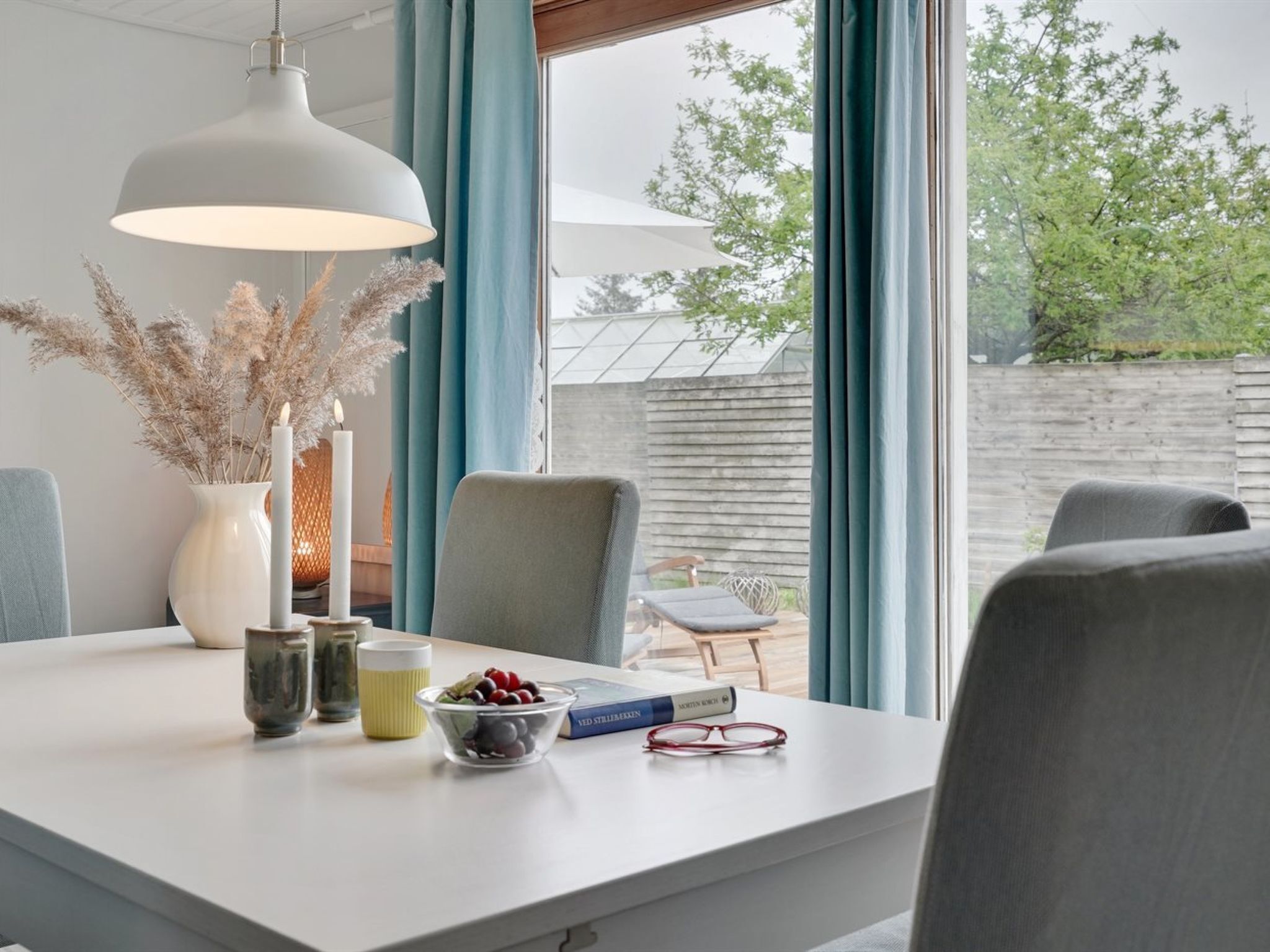 Et moderne spiseområde med et hvidt bord, en hængende lampe og udsigt til det fri via store vinduer.