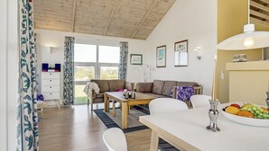 A modern living room with a wooden ceiling, a sofa, a dining table, and a view of the outdoors through large windows.
