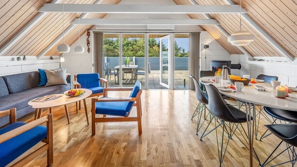 Een ruime zolderkamer met een hoge nok, houten balken, een eethoek met tafel en stoelen, een bank en een grote schuifdeur met toegang tot een terras.