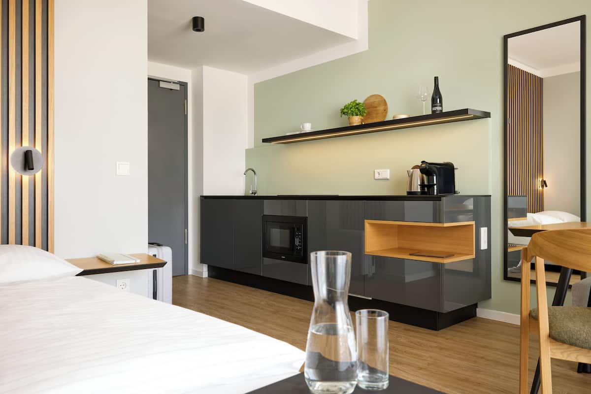 Quarto de hotel moderno com cama, mesa de cabeceira, escrivaninha, cadeira, espelho e área de cozinha com micro-ondas, cafeteira e prateleira com planta e garrafa de vinho.