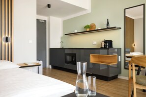 A modern hotel room with a bed, a bedside table, a nightstand, a desk, a chair, a mirror, a kitchen area with a microwave, a coffee maker, and a shelf with a plant and a wine bottle.