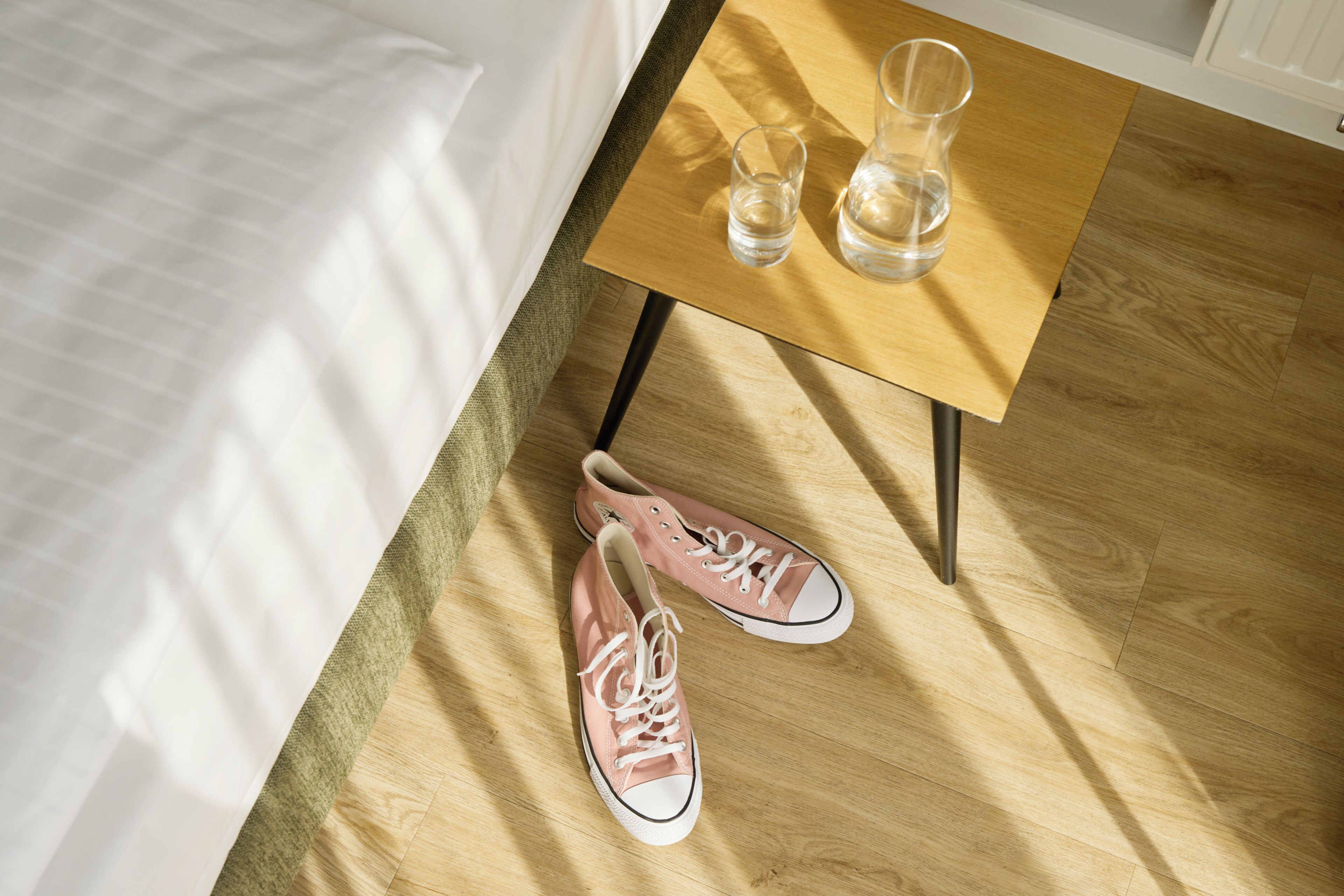 A bed, a wooden table with two glasses, and a pair of pink shoes on a wooden floor.