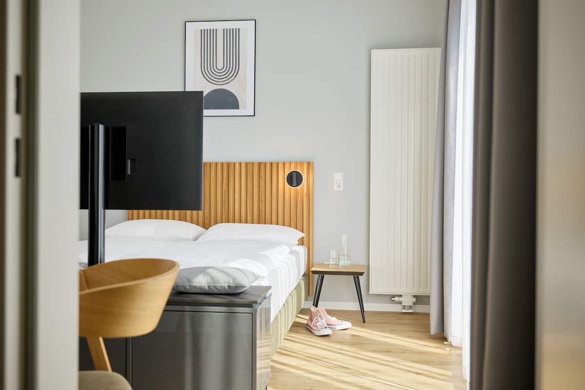Quarto de hotel moderno com cama, cabeceira de madeira, televisão, mesinha com copos e uma cadeira.