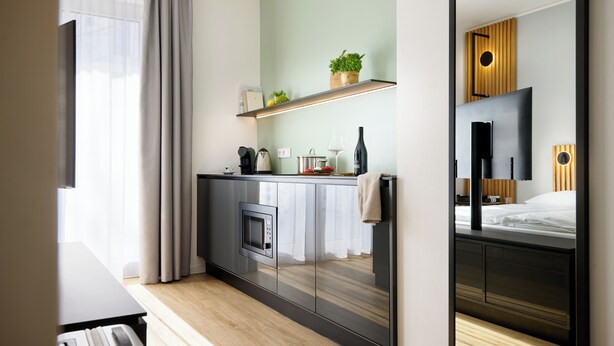 A modern hotel room with a kitchenette, a bed, a large mirror, and a view of the city. - appartello - smarttime living Berlin (Berlin)