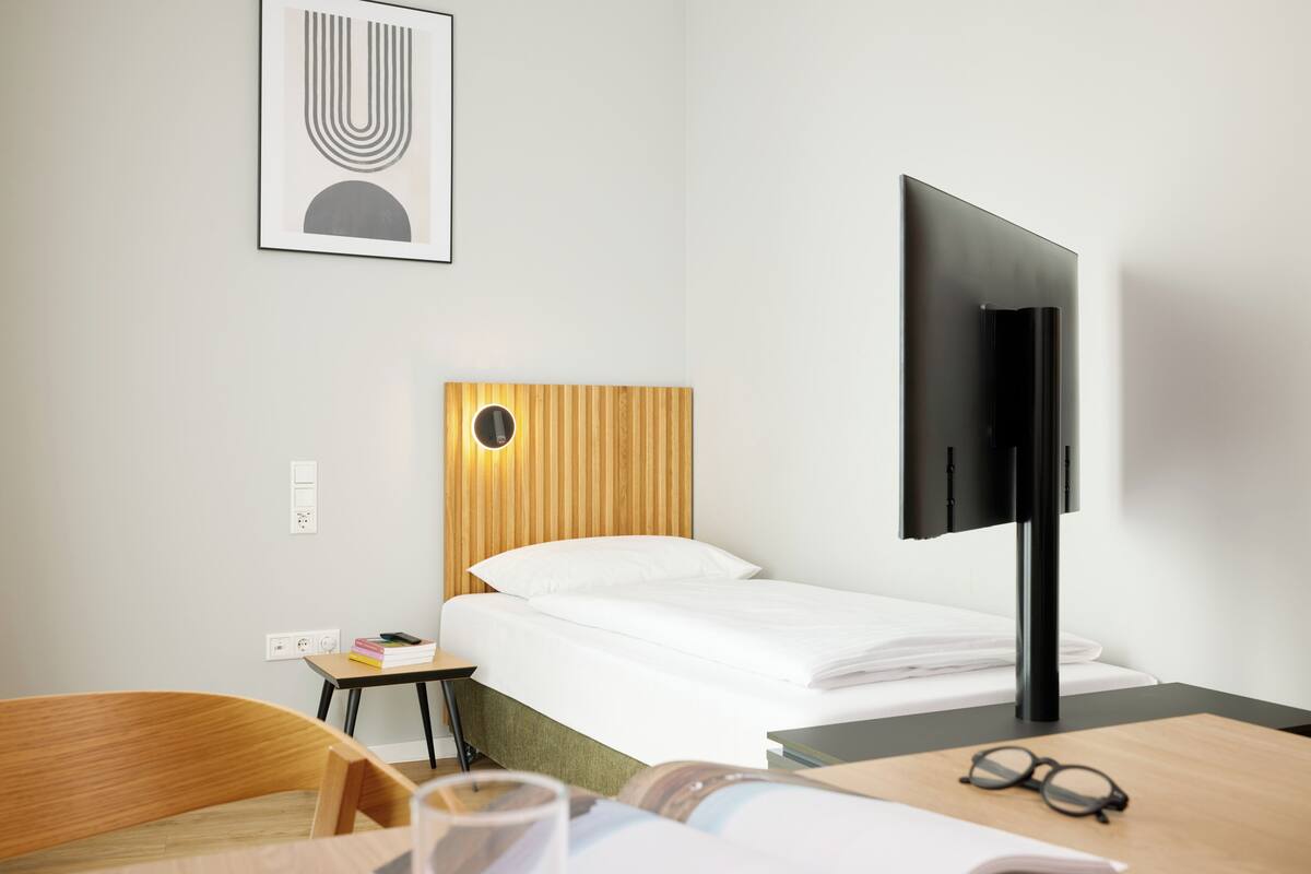 Quarto de hotel com uma cama, uma escrivaninha com cadeira, televisão e um quadro na parede.