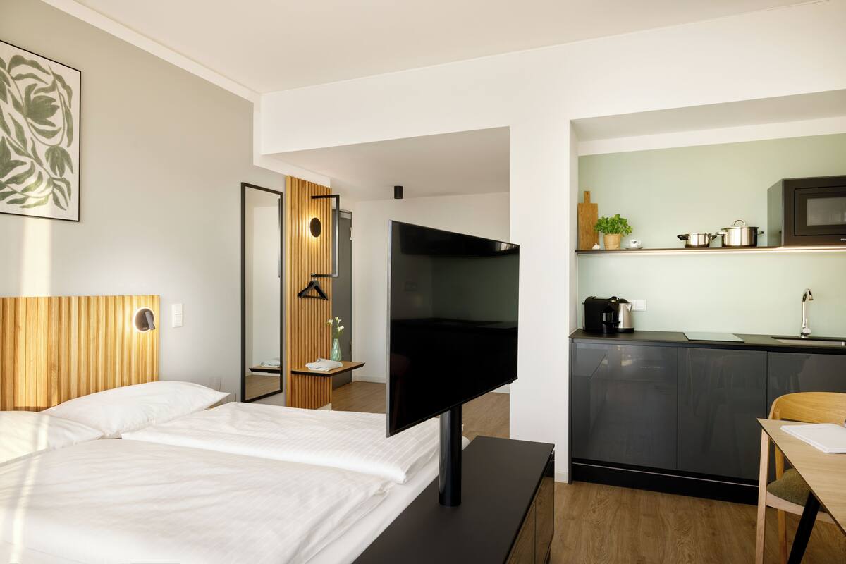 Quarto de hotel moderno com uma cama, televisão, cabeceira de madeira, espelho e pequena área de cozinha com micro-ondas, além de uma mesa de madeira.