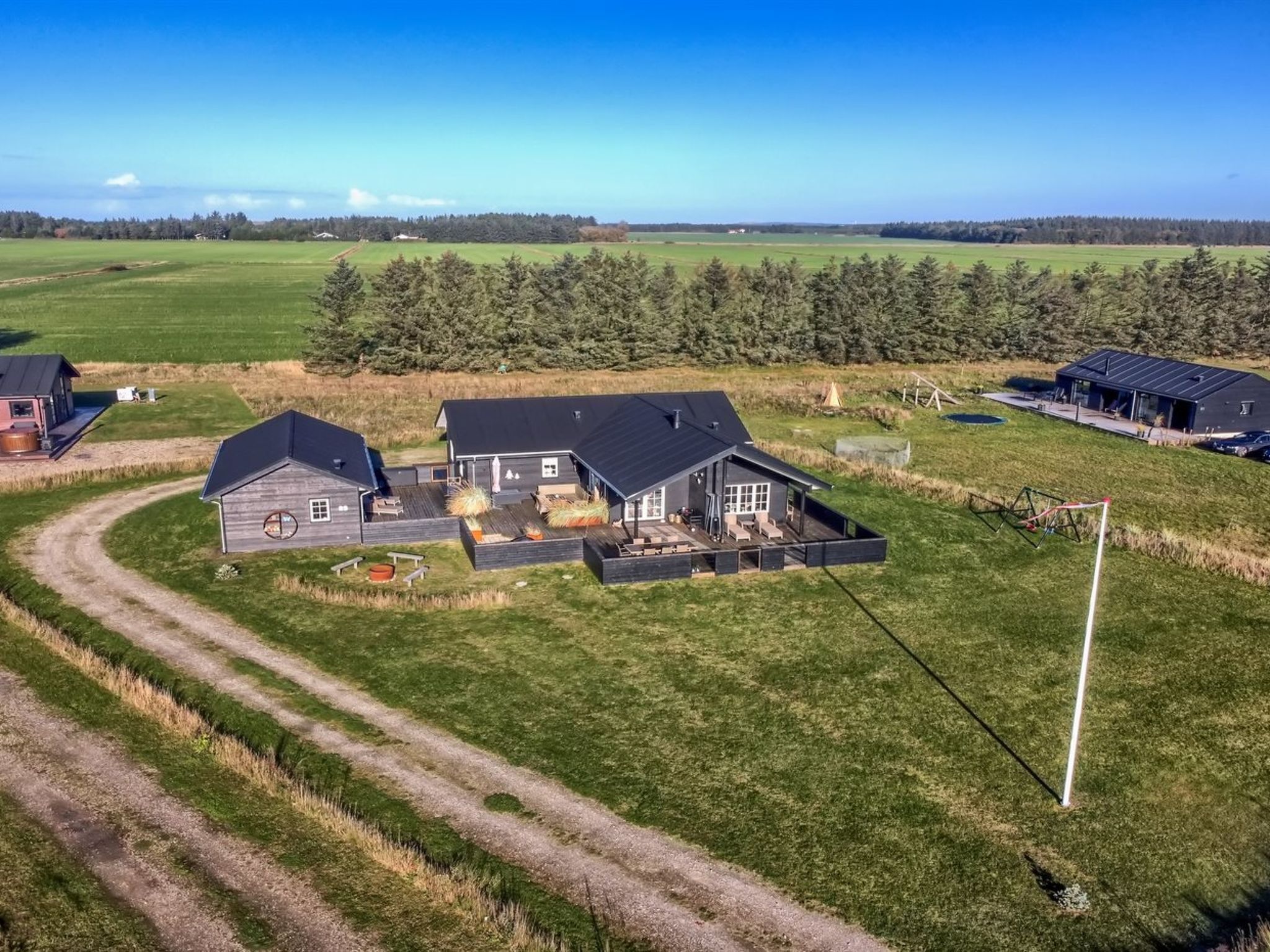 A large, modern house with a spacious patio area, surrounded by open fields and a forest in the background.