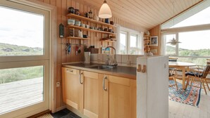 Une cuisine avec des armoires en bois, un évier et un réfrigérateur. Des étagères se trouvent au-dessus du plan de travail, où sont rangés divers ustensiles de cuisine.