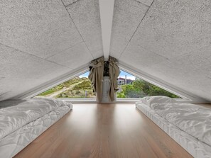 A bedroom with a sloped ceiling, two beds, a window with curtains, and a view of greenery and buildings outside.