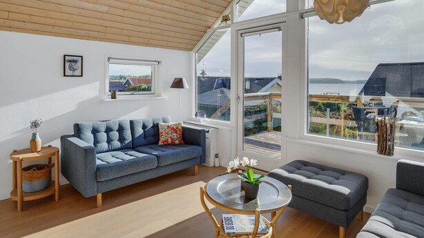 Una sala de estar moderna con un sofá azul, una mesa redonda de cristal y un reposapiés gris. Amplias ventanas ofrecen vistas del entorno.