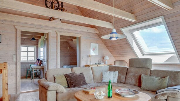 Een knus loft-achtig vertrek met een houten plafond, een groot raam en een bank met kussens.
