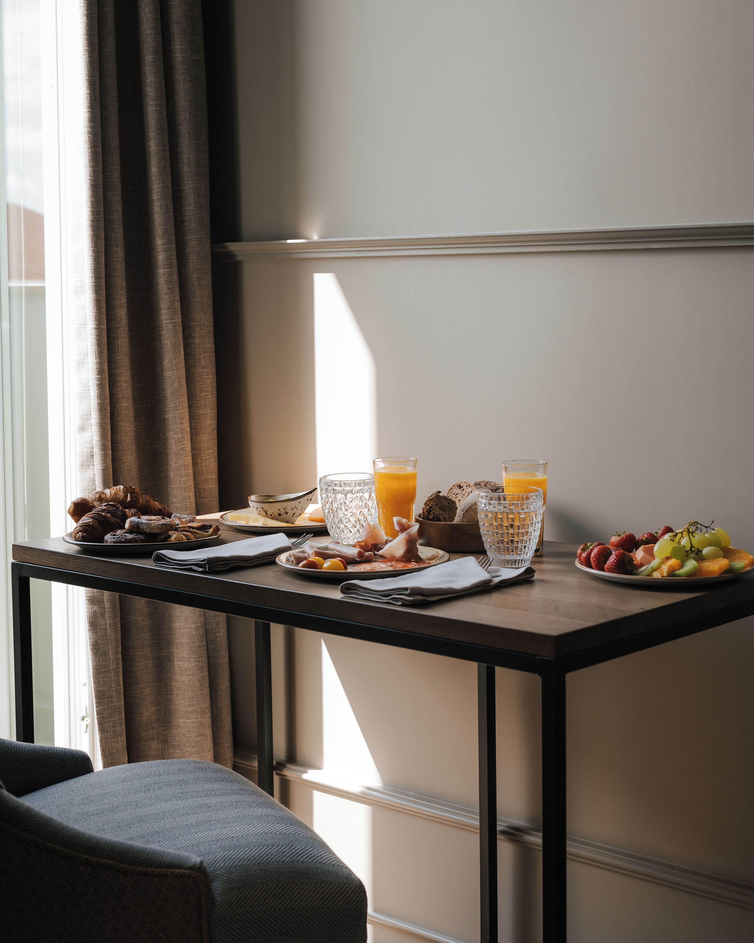 Une table de petit-déjeuner avec des croissants, des fruits et du jus, placée devant une fenêtre ornée de rideaux.