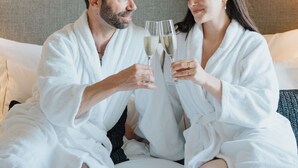 A couple in white bathrobes sitting on a bed with a headboard, holding champagne glasses. - AlmaLusa Alfama (Lisbon)
