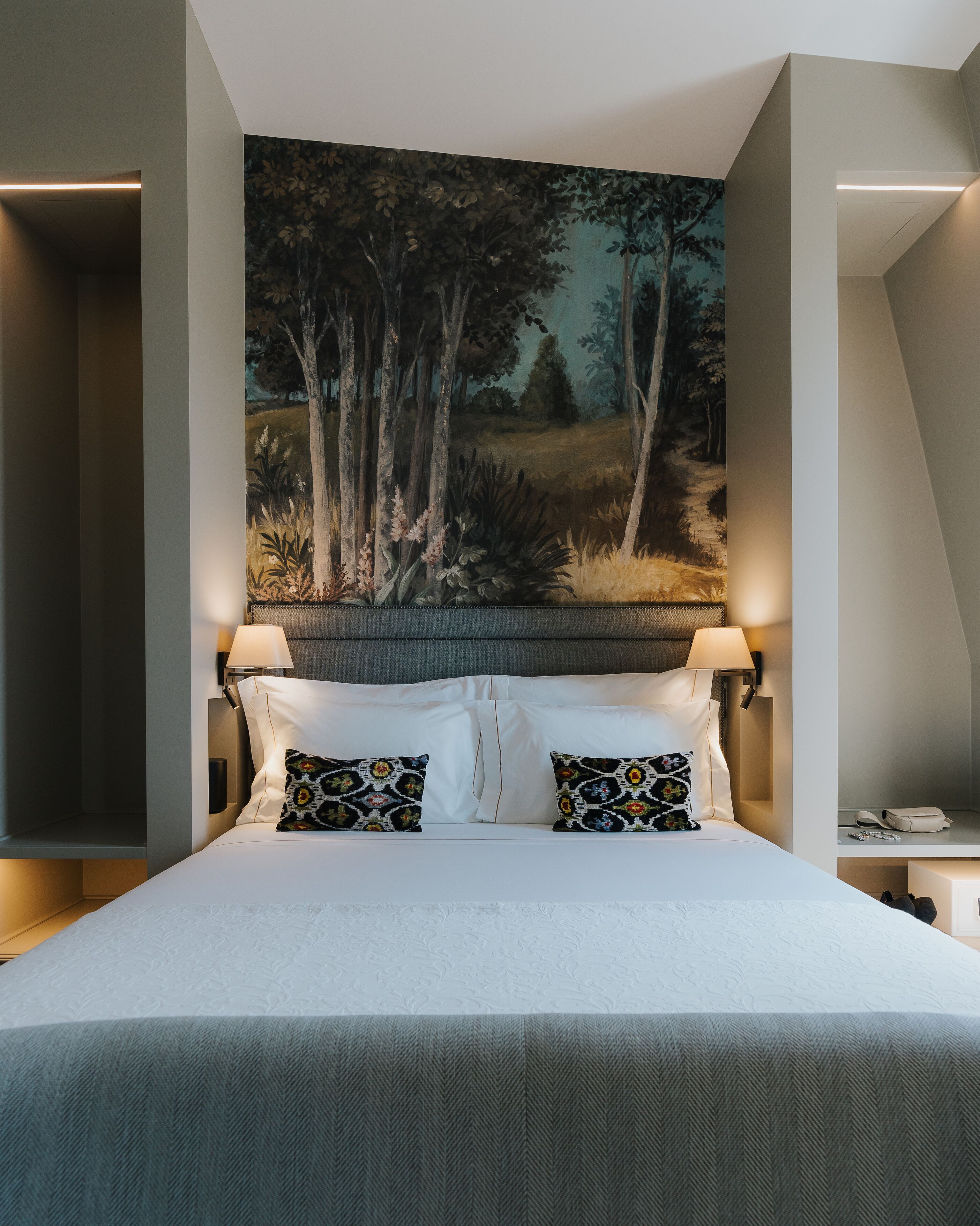A hotel room with a large bed, two wall-mounted lamps, a painting of a forest, and a small bedside table with a lamp.