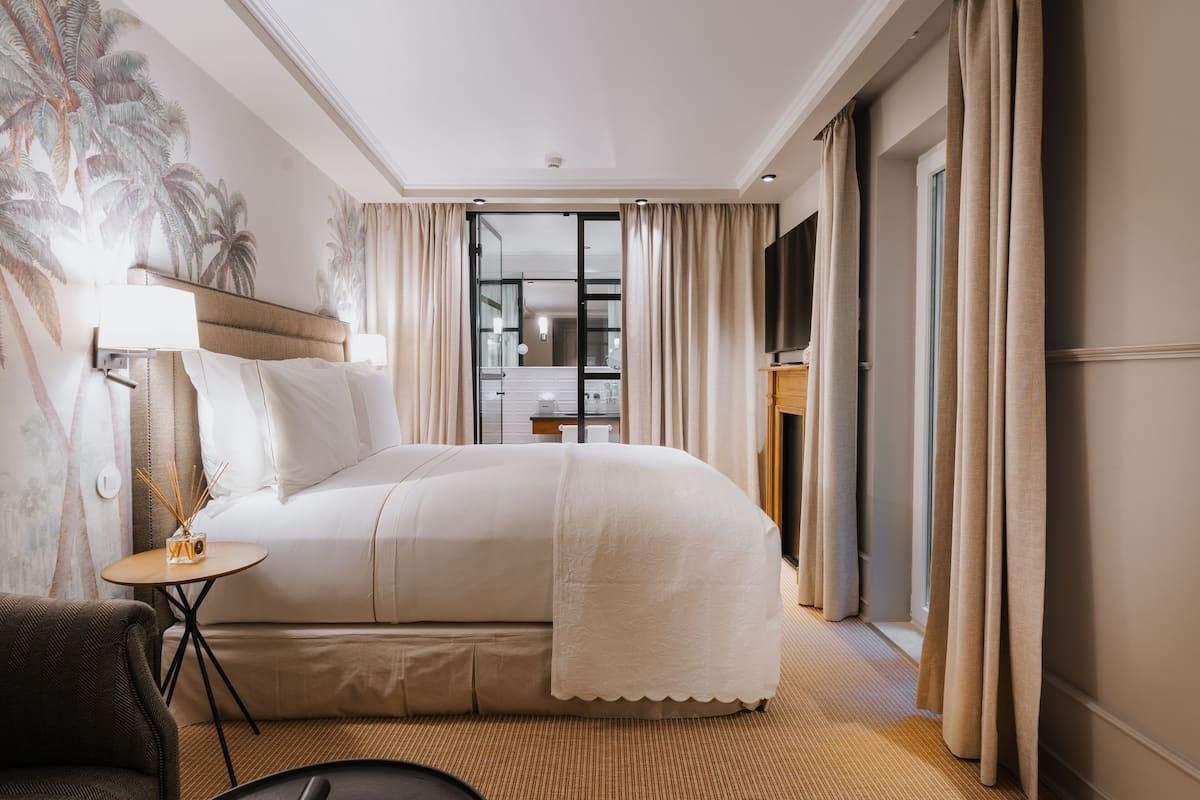 Hotelkamer met een groot bed, een klein tafeltje met een lamp, een stoel en uitzicht op de badkamer via een glazen deur.