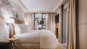 A hotel room with a large bed, a small table with a lamp, a chair, and a view of the bathroom through a glass door.