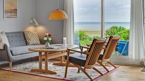 A living room with a sofa, two wooden chairs, a table, and a large window with a view of the beach. - "Elpida" - 100m from the sea by Interhome (Glesborg)