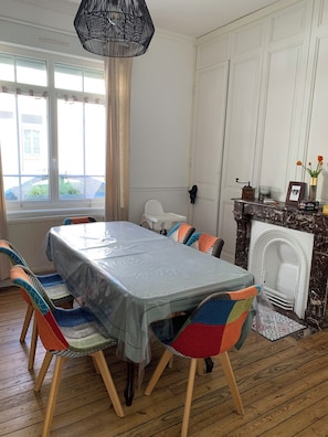 Een eetkamer met een tafel afgedekt met plastic, kleurrijke stoelen, een open haard en een raam met gordijnen.